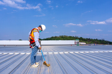 Worker wearing full safety body harness holding a broom in hand  working on roof top for cleaning metal roof sheet