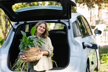 Woman with plant in pot next to a charging electric car in the yard of a country house