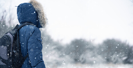 Back view of traveler in a blue jacket with a fur hood and a backpack on the background of a winter...