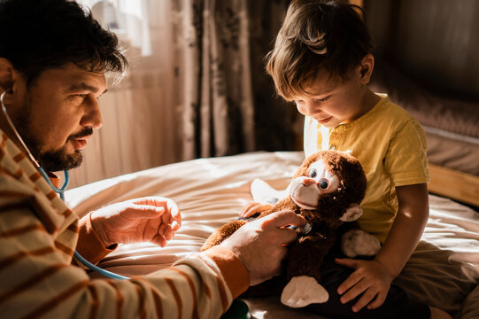 Man examining stuffed toy with stethoscope and son sitting on bed at home