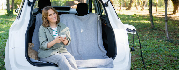 Naklejka premium Woman with smartphone sits in an electric car's trunk