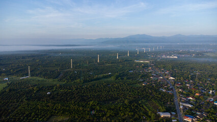 Aerial view of High voltage grid tower with wire cable at tree forest with fog in early morning. Colorful landscape with woods in fog, sunbeams, sky, forest in winter morning.