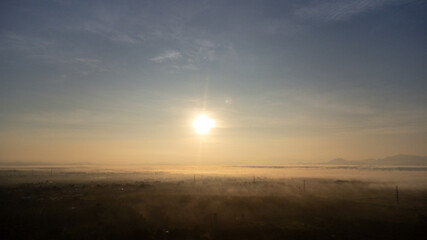 Aerial view of High voltage grid tower with wire cable at tree forest with fog in early morning. Colorful landscape with woods in fog, sunbeams, sky, forest in winter morning.