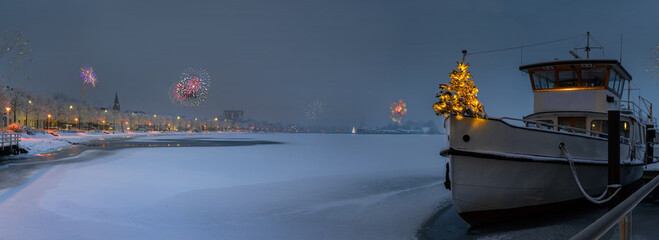 Silvester time on the Schlei. Fireworks on New Year Ferry with Christmas tree standing at edge of bow. Panorama of ship boat moored at the pier covered with snow and promenade in winter in Schleswig. 