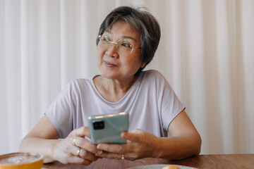 Happy asian old elder woman smiling and looking at empty space, holding and using phone, chatting or reading news on social, sitting on chair alone over white curtain.