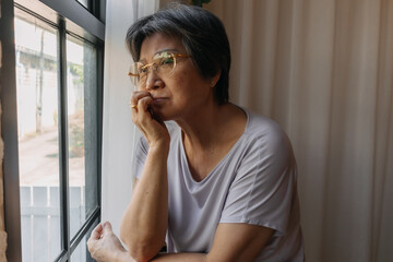 Asian old woman mother sitting on chair and looking out the window, resting chin on arm, missing...