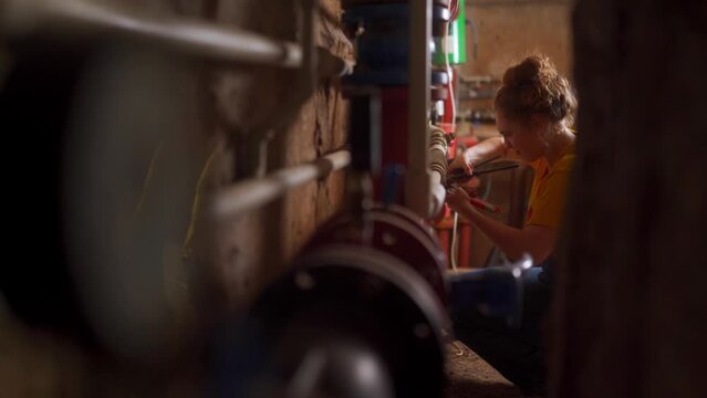 Young professional female plumber fixes water pipes using adjustable and pliers wrenches in bolier room. Side shot of woman carries out repair work with water supply system