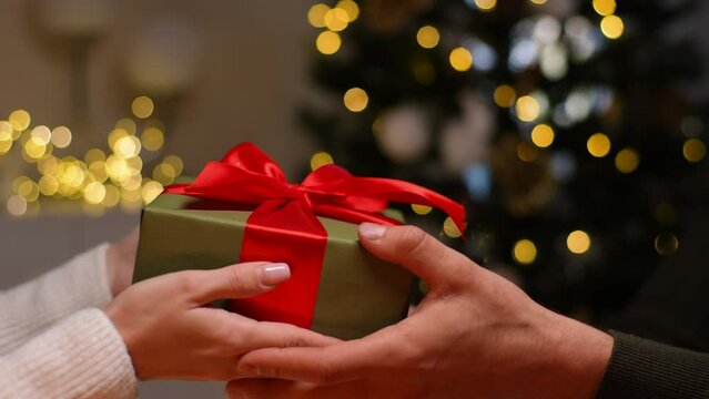 Close-up hands of unrecognizable young woman giving Christmas gift to loving man on background Christmas tree with bright blurred lighting at cozy dark living room, slow motion, side view.