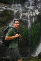 Portrait of handsome male tourist with backpack standing in front of the tropical waterfall