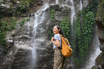Image of smiling female tourist with backpack enjoying the view of beautiful tropical waterfall