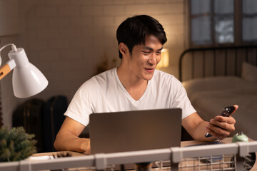 Very happy Asia teenager man use smartphone at night time working with computer notebook in bed room	