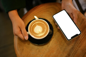 Above view of young woman holding cup of hot latte coffee and using mobile phone