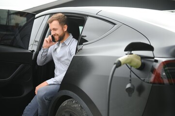 Happy man using smart phone and charging car at vehicle charging station