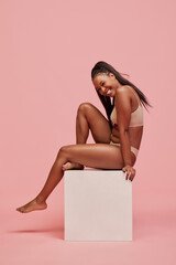 Beautiful, smiling young African woman with long hair posing in underwear against pink studio...