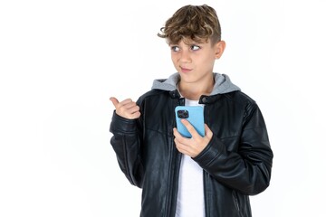  caucasian kid boy wearing black jacket points thumb away and shows blank space aside, holds mobile...