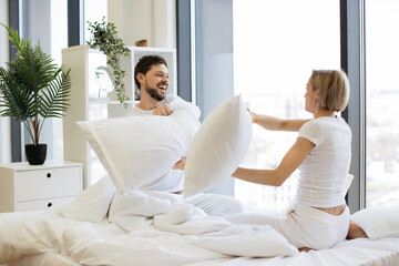Side view of happy loving Caucasian husband and wife couple having pillow fight in bed, happily spending time on weekend.