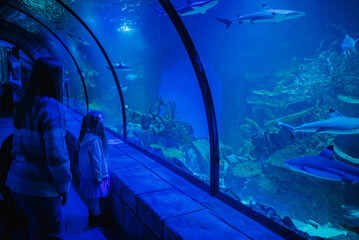 Cute little daughter with mother visiting zoo aquarium. People fascinated observing fish at...