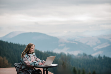 Girl working with a great view mountains. Workplace in country in backyard house in morning. Woman remote work on laptop while sitting at a table outdoors. Concept freelance lifestyle. Internet 5G.