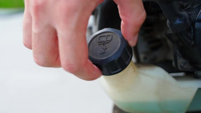 A hand unscrews the fuel tank cap of a garden trimmer, close-up. Blue gasoline oil mixture for two-stroke lawnmower engine. Checking the fuel level before topping up