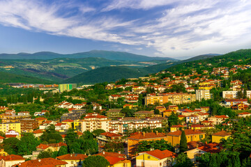 Fototapeta na wymiar panoramic landscape view of green summer city with yellow houses, green forest and mountains above and amazing hills and cloudy sky on background