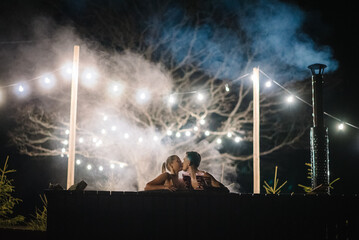 Couple enjoying hot tub at night outdoors. Winter holidays in mountains. Family relax in thermal spa open air. Man and woman swims in hot bath while resting. Glowing garlands and light bulbs. - obrazy, fototapety, plakaty