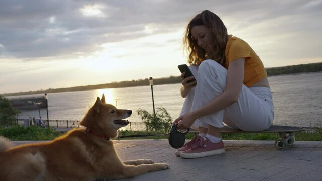 A woman takes pictures of her red-haired dog on a walk for social networks while sitting on a skateboard in the evening at sunset