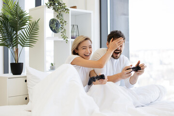 Beautiful young woman sitting in bed and cheating closing eyes of husband while playing video games...