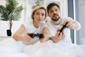 Close up of concentrated caucasian couple playing video games while lying under blanket at home. Smiling man and woman looking on tv have fun competing in video game using joysticks.