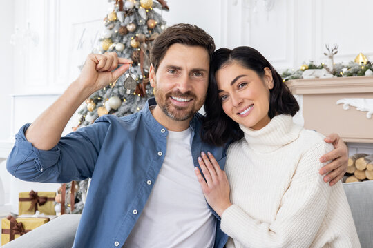 Portrait of a young family, a man and a woman sit on the sofa in a new apartment, smile, cuddle and look at the camera, hold the keys to the house, celebrate the New Year and Christmas