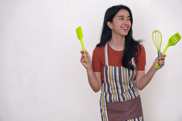 Young Asian woman standing with a big smile while her hands holding some kitchen utensils