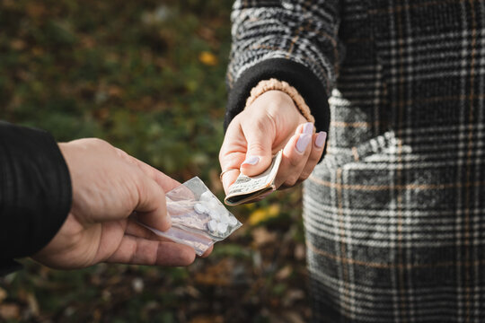 Male drug dealer sells hard drugs in a transparent plastic bag to an addicted woman with money in her hand, cropped image
