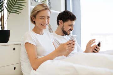 Serene caucasian family of two using mobile phones while lazing in soft bed during daytime at home. Confident adult man and woman reading online article paying attention to details.