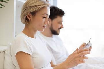 Happy caucasian couple holding cellphones in hands while lounging in soft bed all morning indoors. Attractive man and woman browsing internet using mobile application in digital devices.