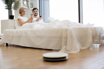 Lovely caucasian couple lying together in comfy bed and using modern laptop while robot vacuum cleaning wooden floor at home. Domestic chores with modern devices.