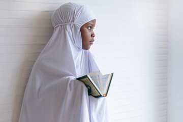 African Muslim child girl wears hijab abaya and holding the Quran, lean against the white wall and...
