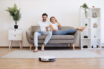 Obraz premium Young caucasian couple in casual clothes relaxing on comfy couch with portable laptop in hands. Family of two watching movie while wireless robot vacuuming wooden floor.