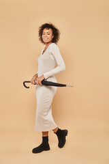 joyful african american woman in midi dress and boots standing with umbrella on beige, autumn style