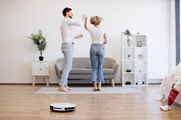 Young caucasian lady dancing with male partner while robotic vacuum running floor smoothly....