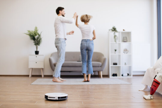 Young caucasian lady dancing with male partner while robotic vacuum running floor smoothly. Homeowners using electrical appliance for household chores while freeing up time for mother-daughter party.