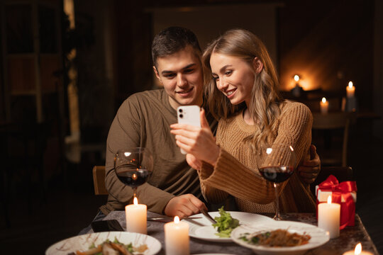 Browsing internet during romantic date dinner at home. Happy couple looking photos on smartphone during celebrating Valentines Day