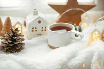 Fototapeta na wymiar Winter hygge. Stylish cup of tea with modern cute christmas houses, pine cone, wooden star and tree, golden lights on soft warm blanket on windowsill. Christmas cozy still life. Merry Christmas!