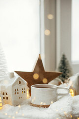 Cozy winter still life. Stylish cup of tea with modern christmas decoration, pine cone, wooden star and tree, golden lights on soft warm blanket on windowsill. Merry Christmas and Happy holidays