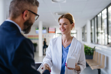 Pharmaceutical sales representative shaking hand with female doctor in medical building. Hospital director consulting with healthcare staff. - obrazy, fototapety, plakaty