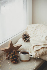 Cozy winter still life. Stylish cup of tea with cozy knitted sweater, pine cone, wooden star and golden lights on windowsill in festive scandinavian room. Autumn and christmas holidays