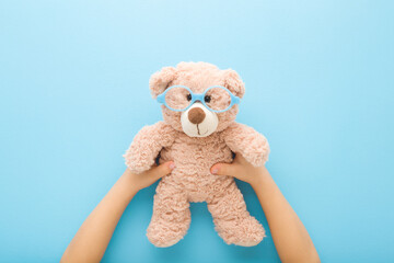 Baby boy hands holding brown teddy bear with eyeglasses with round frame on light blue table background. Pastel color. Closeup. Point of view shot. Children vision concept. Top down view.