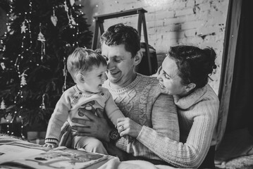 Mother and father reading book for child. Mom and Dad hug son enjoying time together. Young family near Christmas tree at home. Merry Christmas and Happy Holidays. Black and white photo. Closeup.