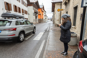 Female tourist in winter black clothes standing on sidewalk looking at his phone while cars ride on road; Venas di Cadore village; Dolomites; Italy