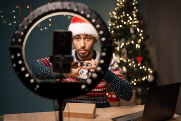 Arab blogger unboxing new smartphone and recording video on camera on Christmas at home. Young guy in Santa hat sharing with subscribers his feedback about modern gadget. - obrazy, fototapety, plakaty