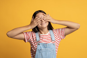 Happy teen girl closing eyes with hands posing on yellow background playing hide and seek game. Teenager brunette female enjoying pastime, feel positive emotions. Advertisement banner poster concept. 