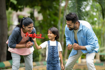 Tracking shot of excited Indian Parents running with girl kid while kid playing with toy airplane at park - concept of Active parenting, Family Bonding and Encouragement.